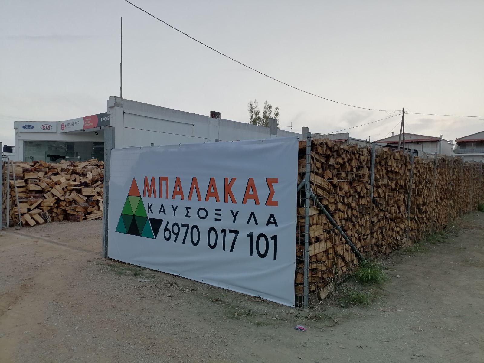 Ταμπέλα Καυσόξυλα Μπαλάκας δίπλα σε στοιβαγμένα ξύλα